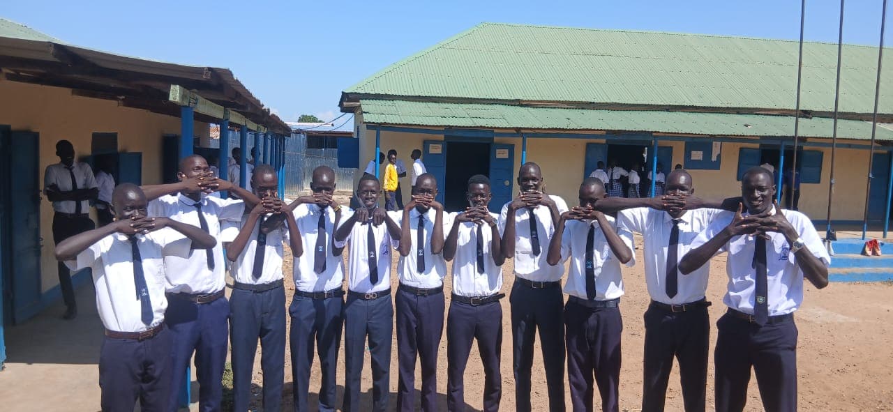 A group of male students in school uniform standing in front of classroom buildings at Gideon Seed Academy Secondary School