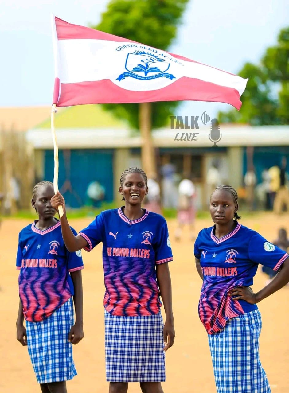 Three students in sports uniform pose together, one holding up the Gideon Seed Academy school flag bearing the school crest