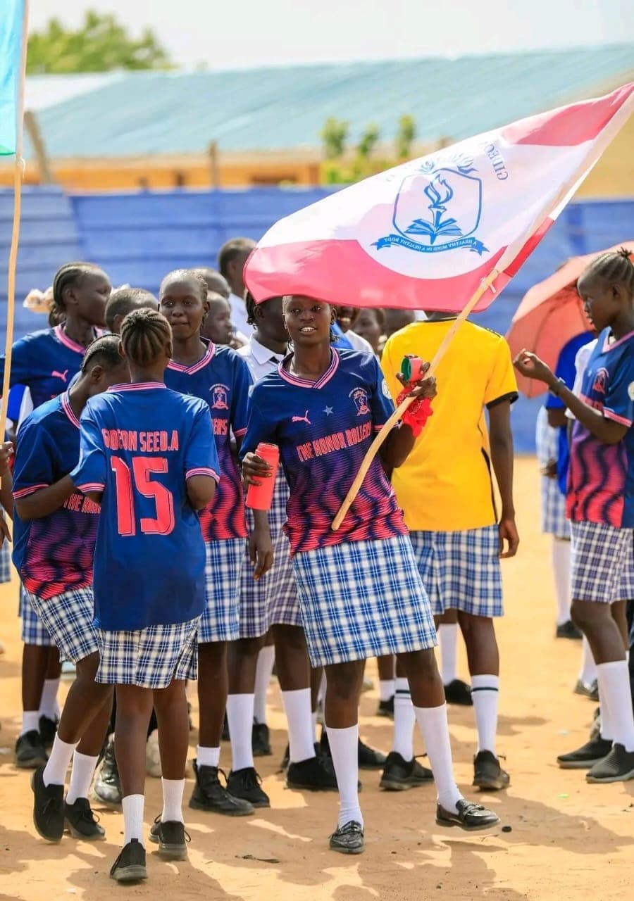 A group of students in sports uniform walk together, one holding the Gideon Seed Academy school flag, with stadium seating in the background